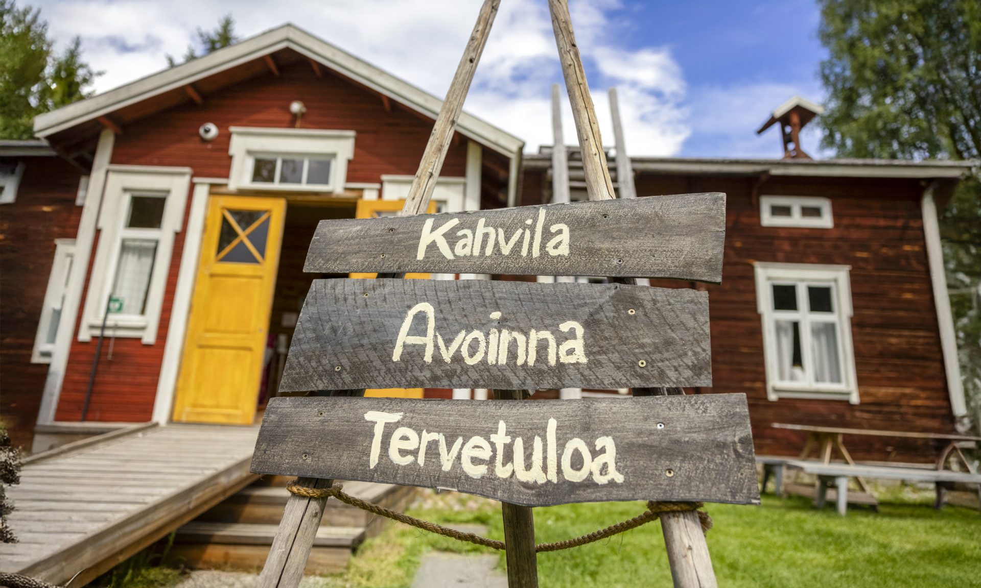 A wooden sign reading 'Café Open, Welcome,' with the Yli-Kärppä house and blue sky in the background.