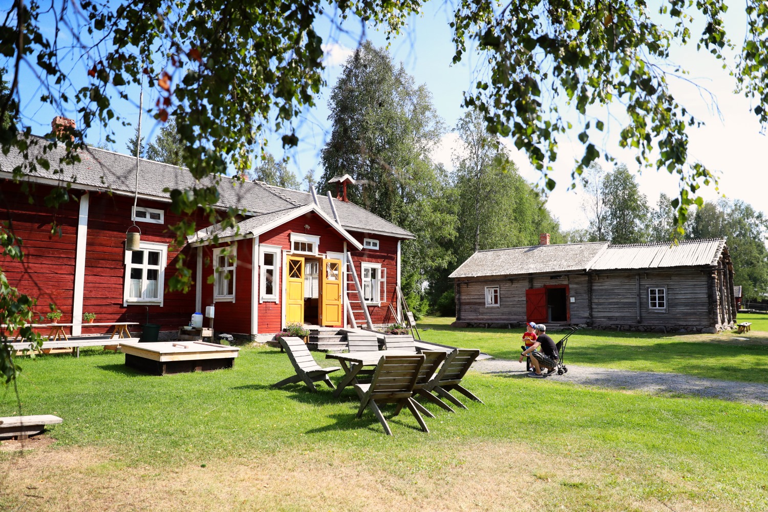 Punainen puinen rakennus, jossa on keltaiset ovet. Rakennuksen edessä puiset pöytäkalusteet.