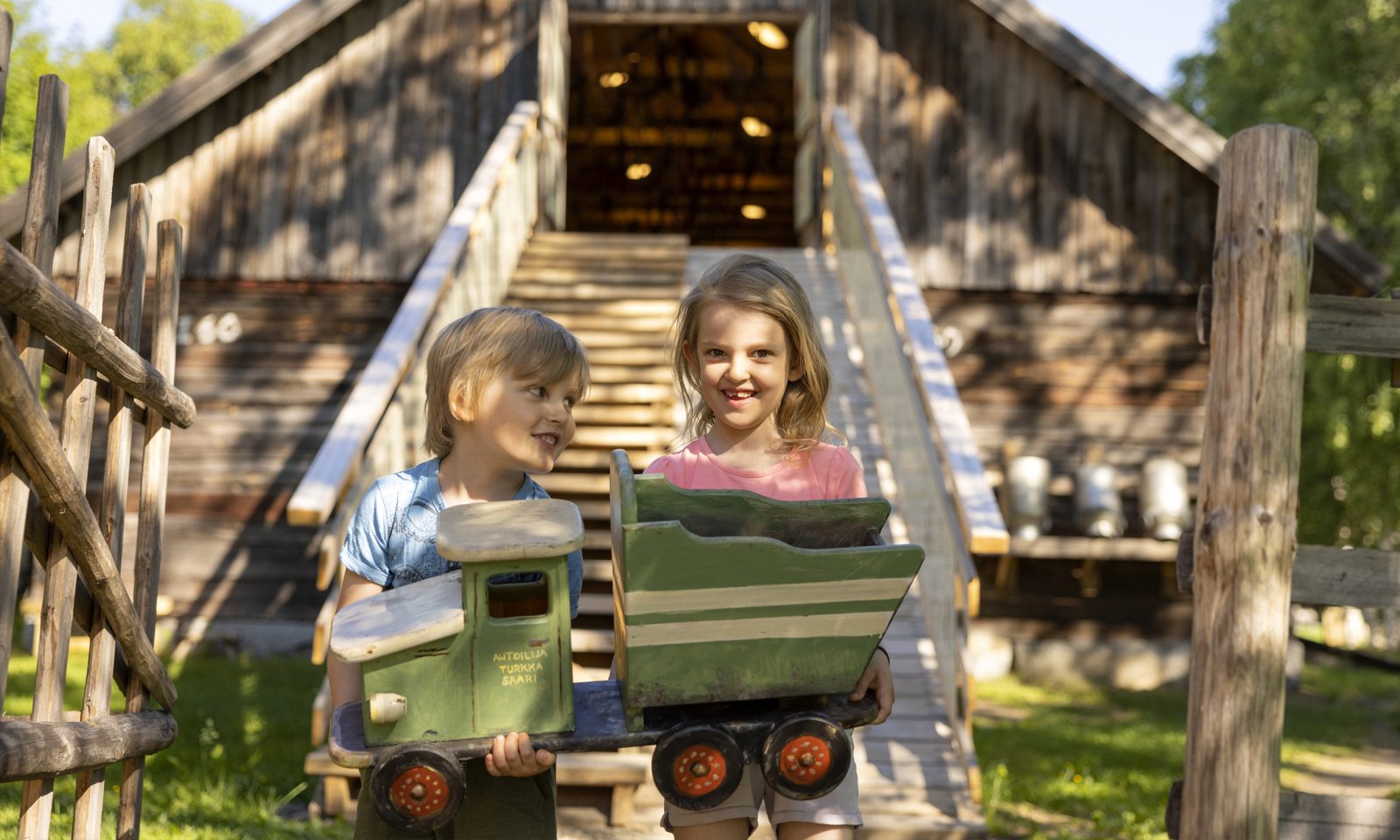 Two children carry a wooden toy truck outdoors on a summer day, with an old wooden granary building and wooden stairs in the background.