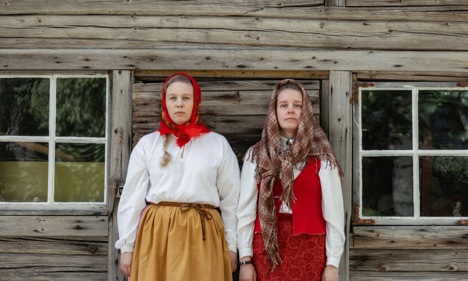 Two women dressed in traditional costumes stand side by side against the wall of an old log building, both with serious expressions on their faces.