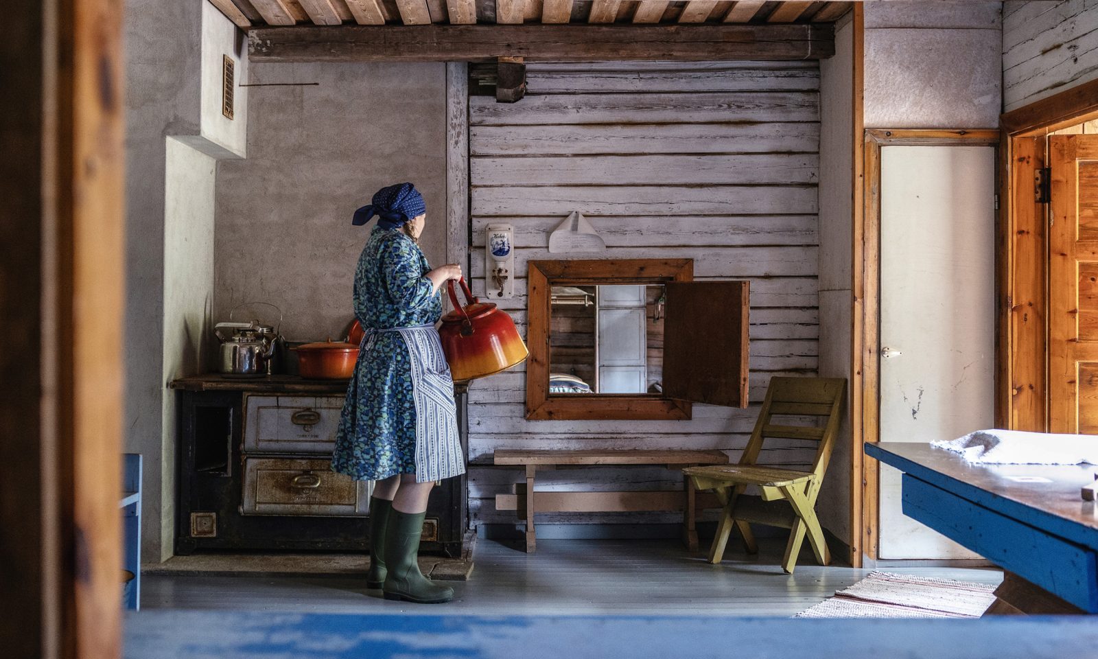 A woman dressed in traditional clothing stands beside a wood-burning stove in an old kitchen, holding a large coffee pot, with log walls and a serving hatch in the background.