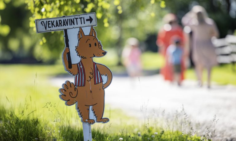 Piirretty kettuhahmo opastekyltissä, jossa lukee "Vekaravintti", taustalla ihmisiä kävelemässä kesäisessä Turkansaaressa.