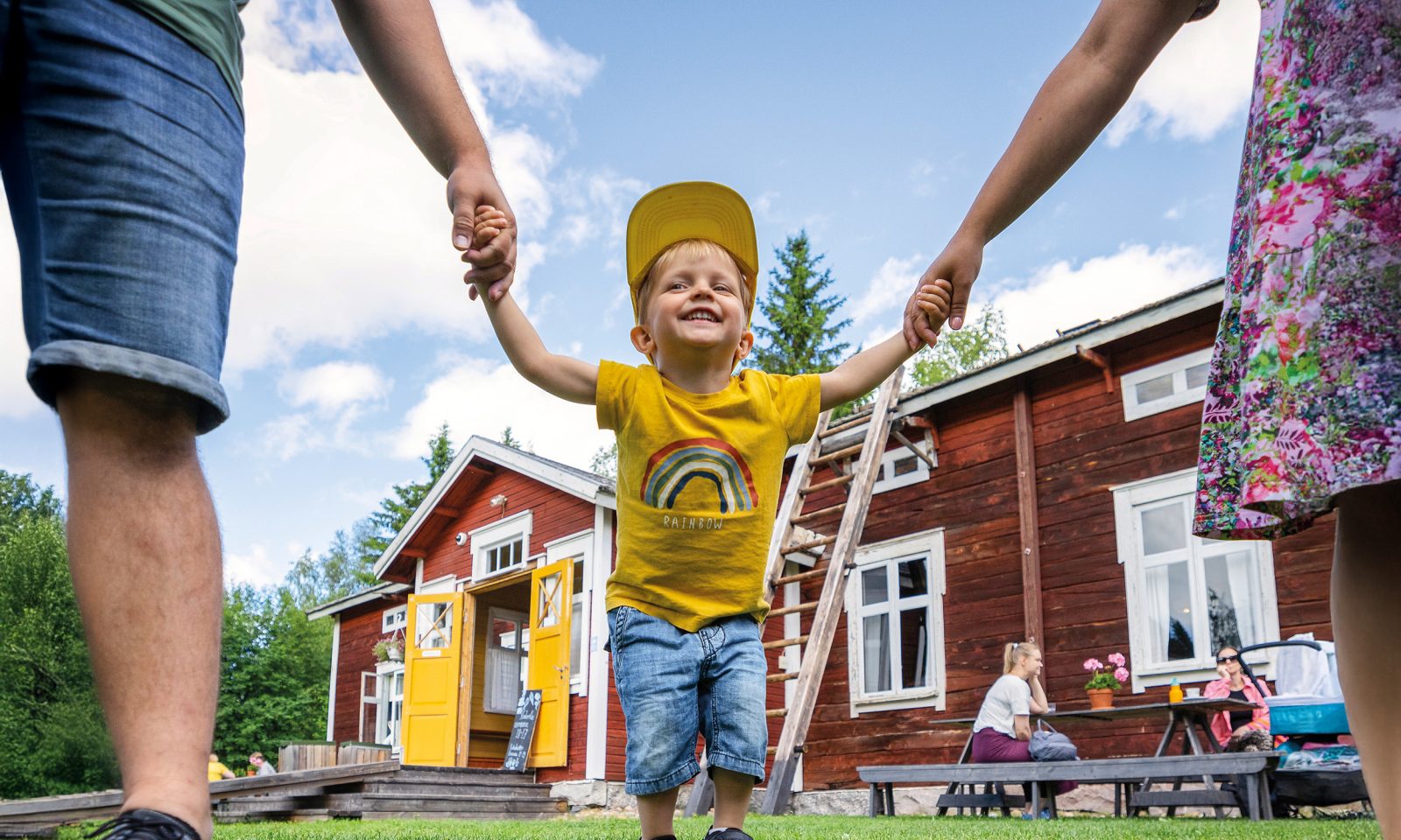 Smiling little boy walks on the lawn in front of a wooden house and holds his parents' hands