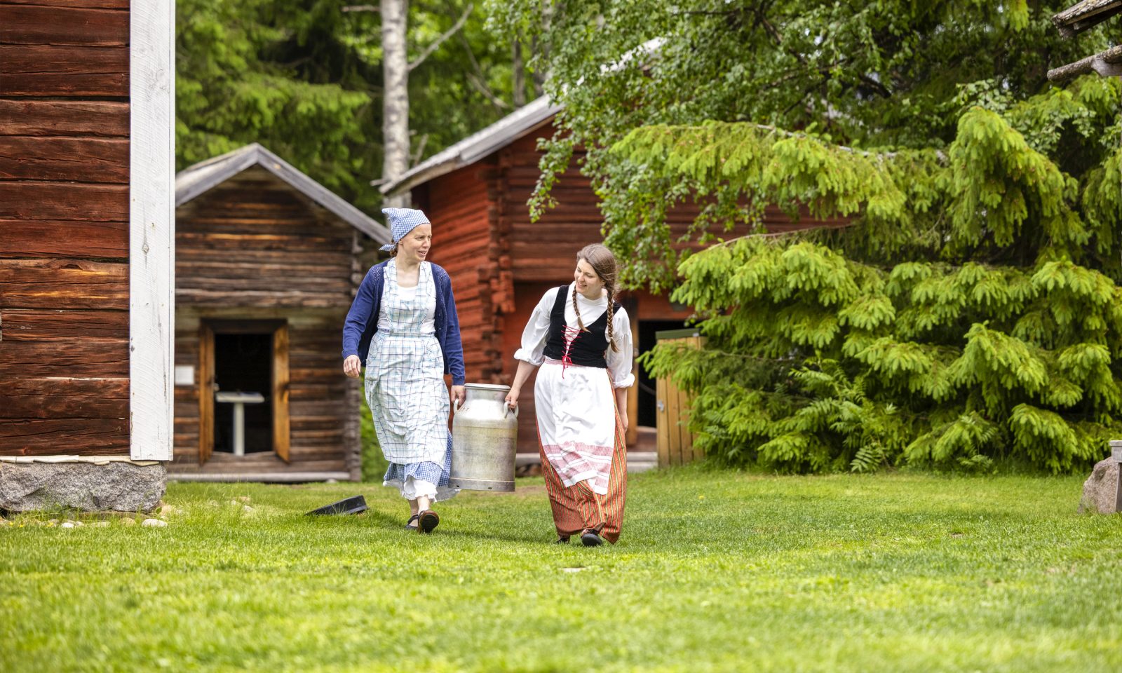 Kaksi perinneasuihin pukeutunutta naista kantaa maitotonkkaa vanhojen hirsirakennusten pihapiirissä.