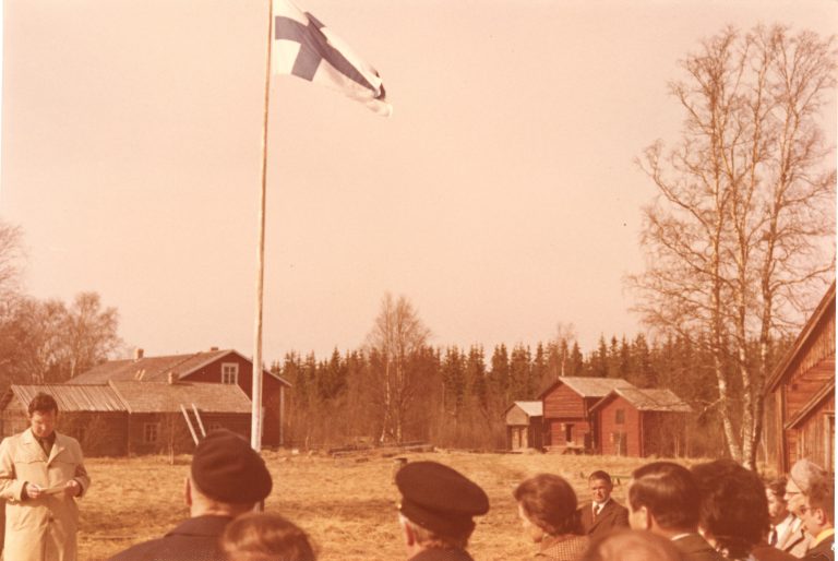 Ihmisiä ja suomen lippu. Taka-alalla vanhoja rakennuksia. Vanha värivalokuva.