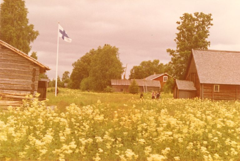 Kukkaniittä, vanhoja rakennuksia ja suomen lippu. Vanha värivalokuva.