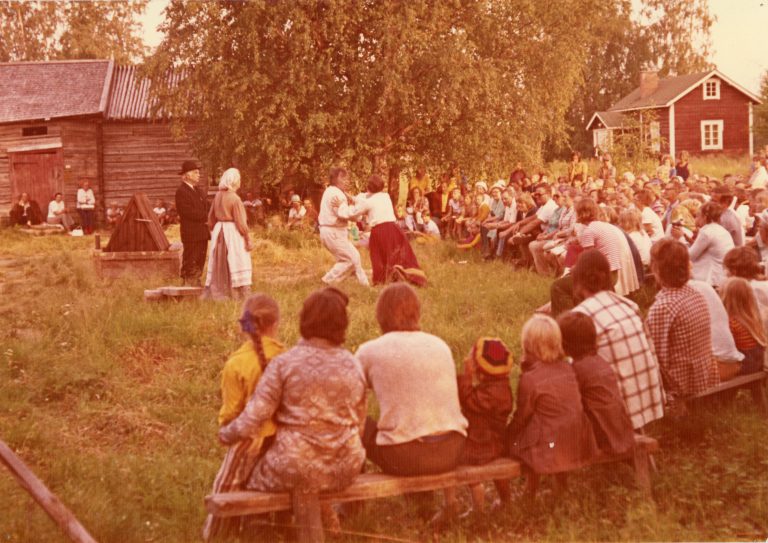 Näyttelijöitä kesäteatterissa sekä puisilla penkeillä istuvia ihmisiä katselemassa esitystä. Taka-alalla vanhoja rakennuksia. Vanha värivalokuva.