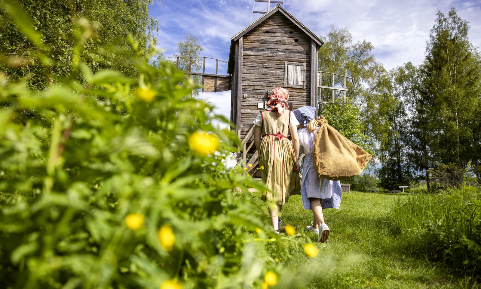 Two girls dressed in traditional clothing walk toward an old windmill in a summery rural landscape. One of them carries a sack of grain on her shoulder.
