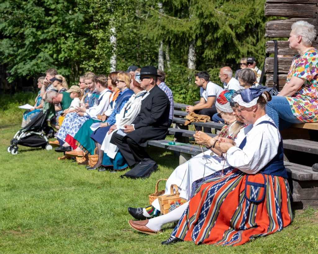 Perinneasuihin pukeutuneita ihimisiä istuu puupenkeillä. Etualalla kaksi naista myös neulovat.