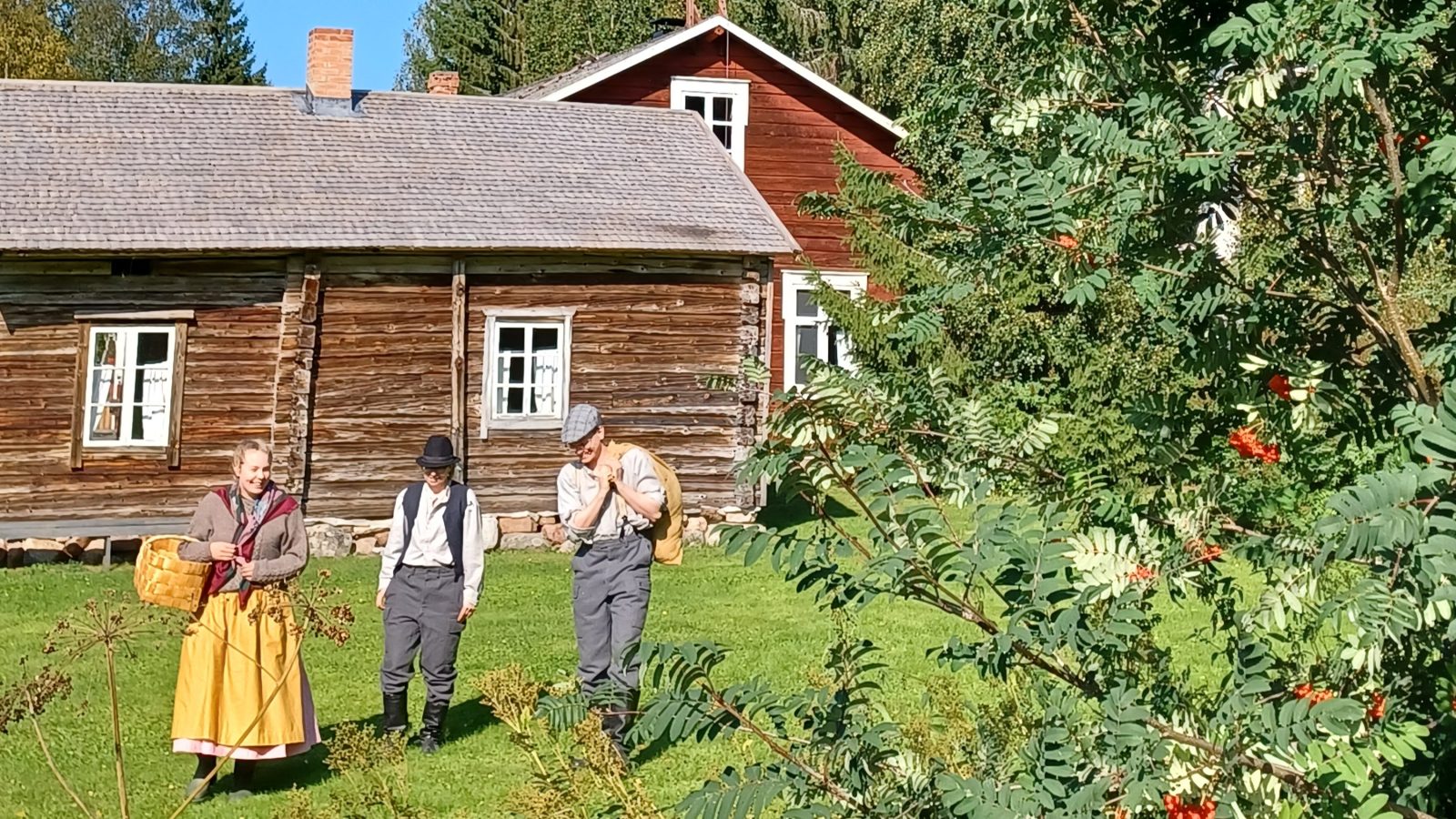 Kolme ihmistä perinneasuissa astelee vanhan rakennuksen pihalla. Etualalla pihlaja marjoineen.